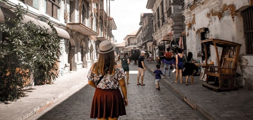 woman in a hat and floral top walking through a cobblestone street in a historic town, surrounded by old buildings and market stalls