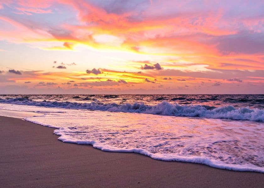 Ocean waves rolling onto a sandy beach beneath a colorful pink, orange, and purple sunset sky.